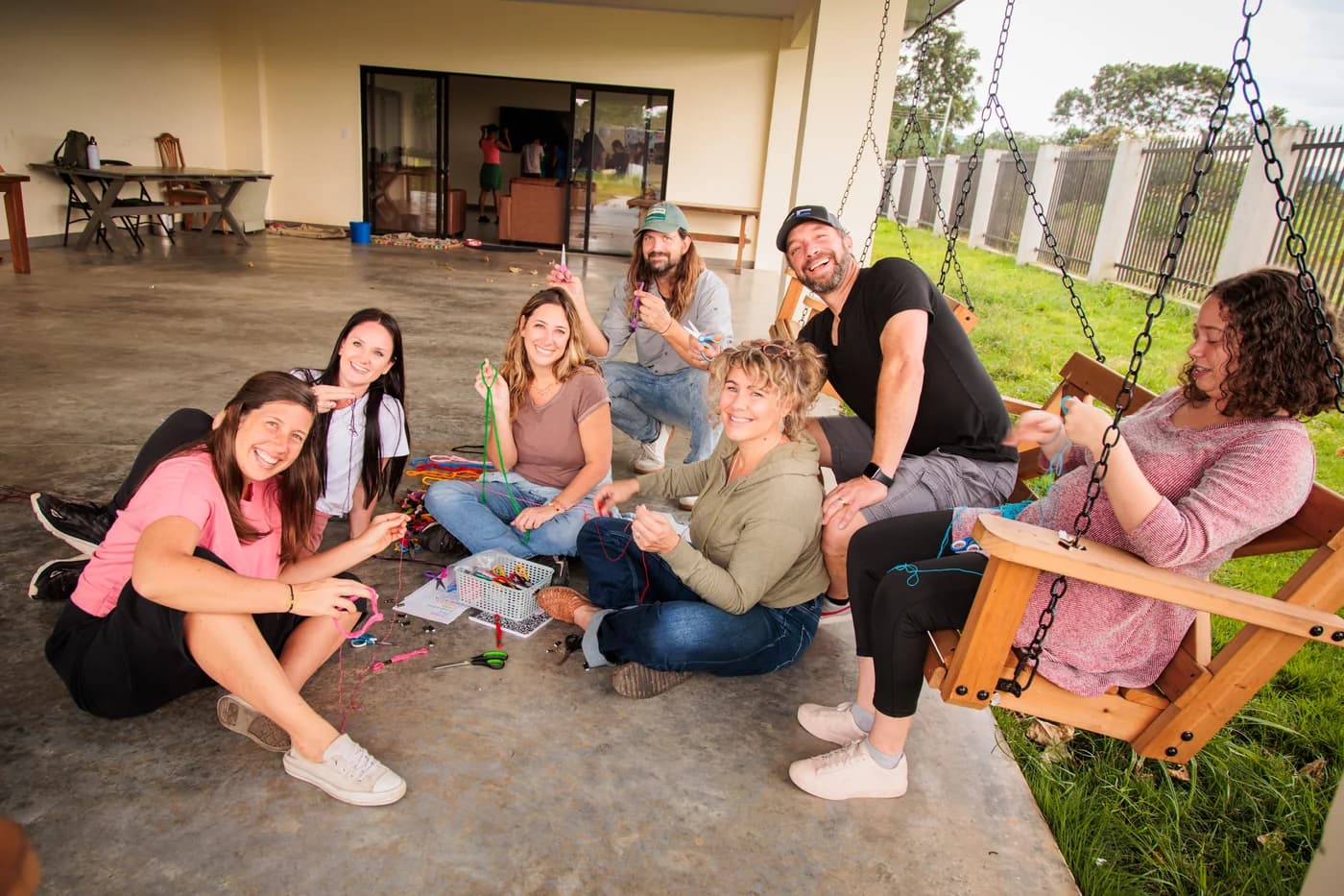 A group sitting together on the floor of an open-sided room in Costa Rica, threading bracelets from a basket of coloured cord, two men among them, everyone looking up and smiling.