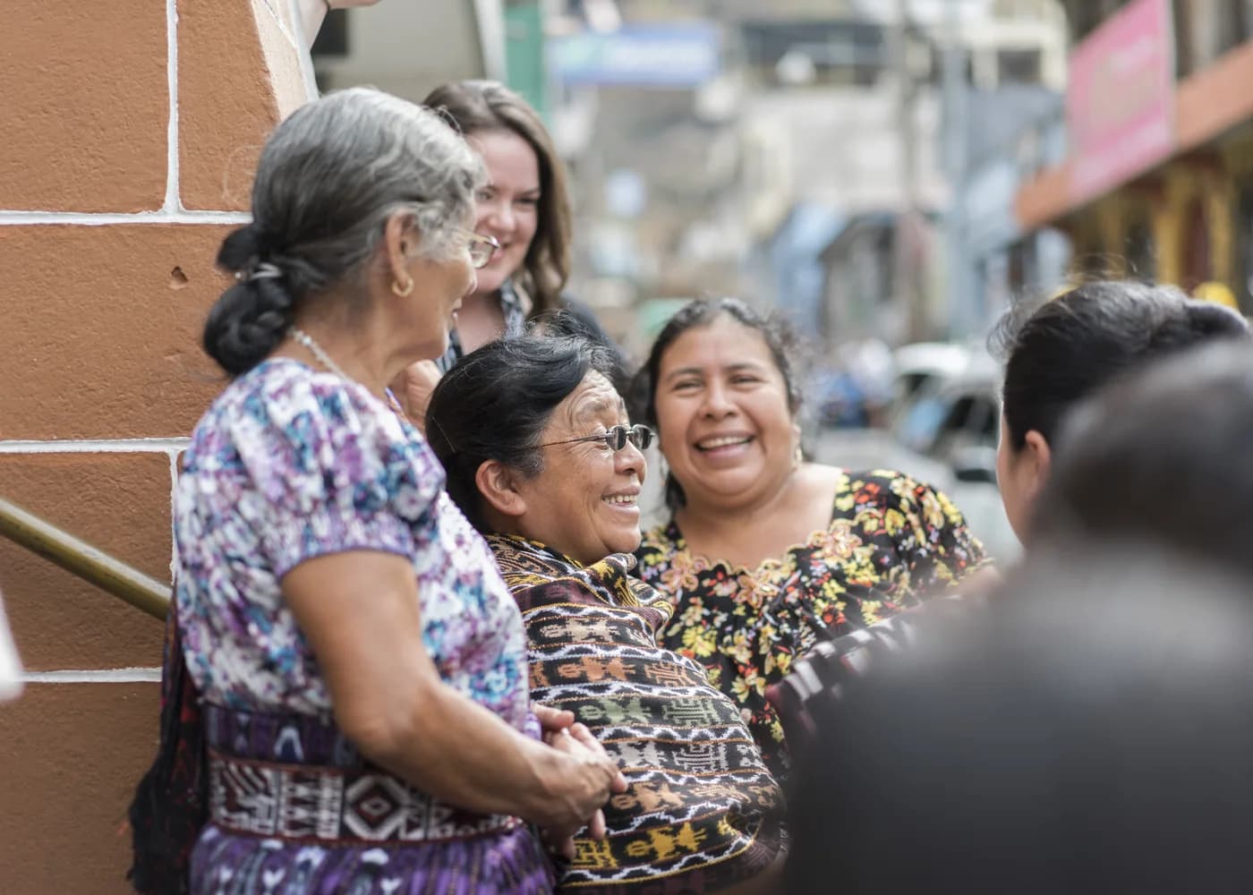 Three women in embroidered blouses laughing together on a street, one of them mid-embrace with a member of the team.