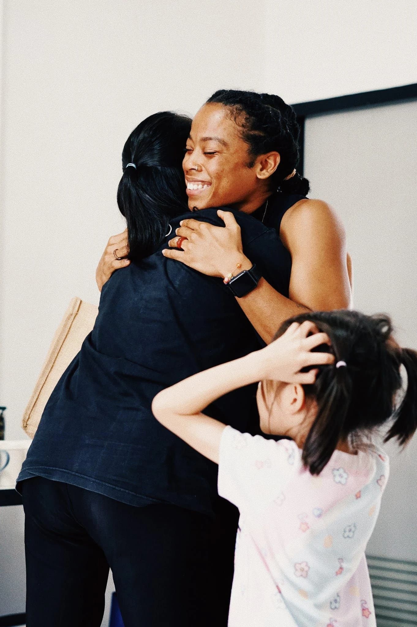 Two women holding each other tightly at the end of a session, one laughing over the other's shoulder, while a small girl reaches up to join the hug.