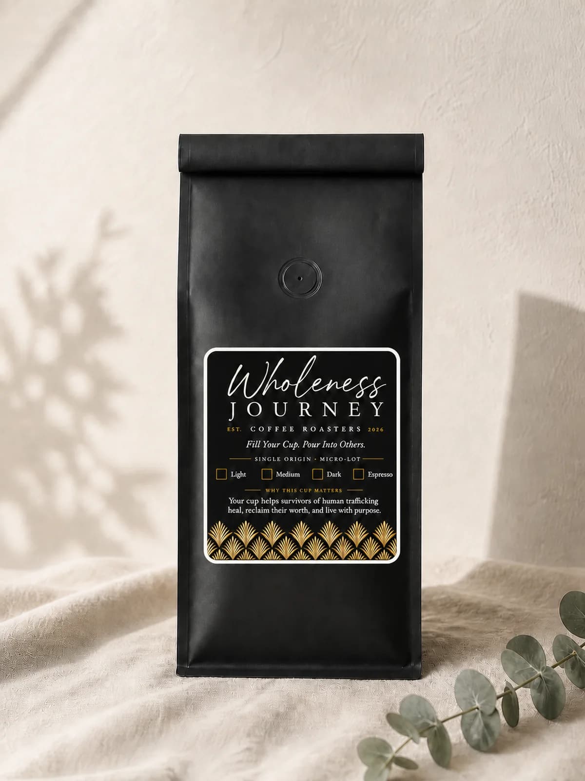 A matte black Wholeness Journey Coffee Roasters bag standing on linen against a plaster wall, its label black with gold fan detail, a eucalyptus sprig beside it.