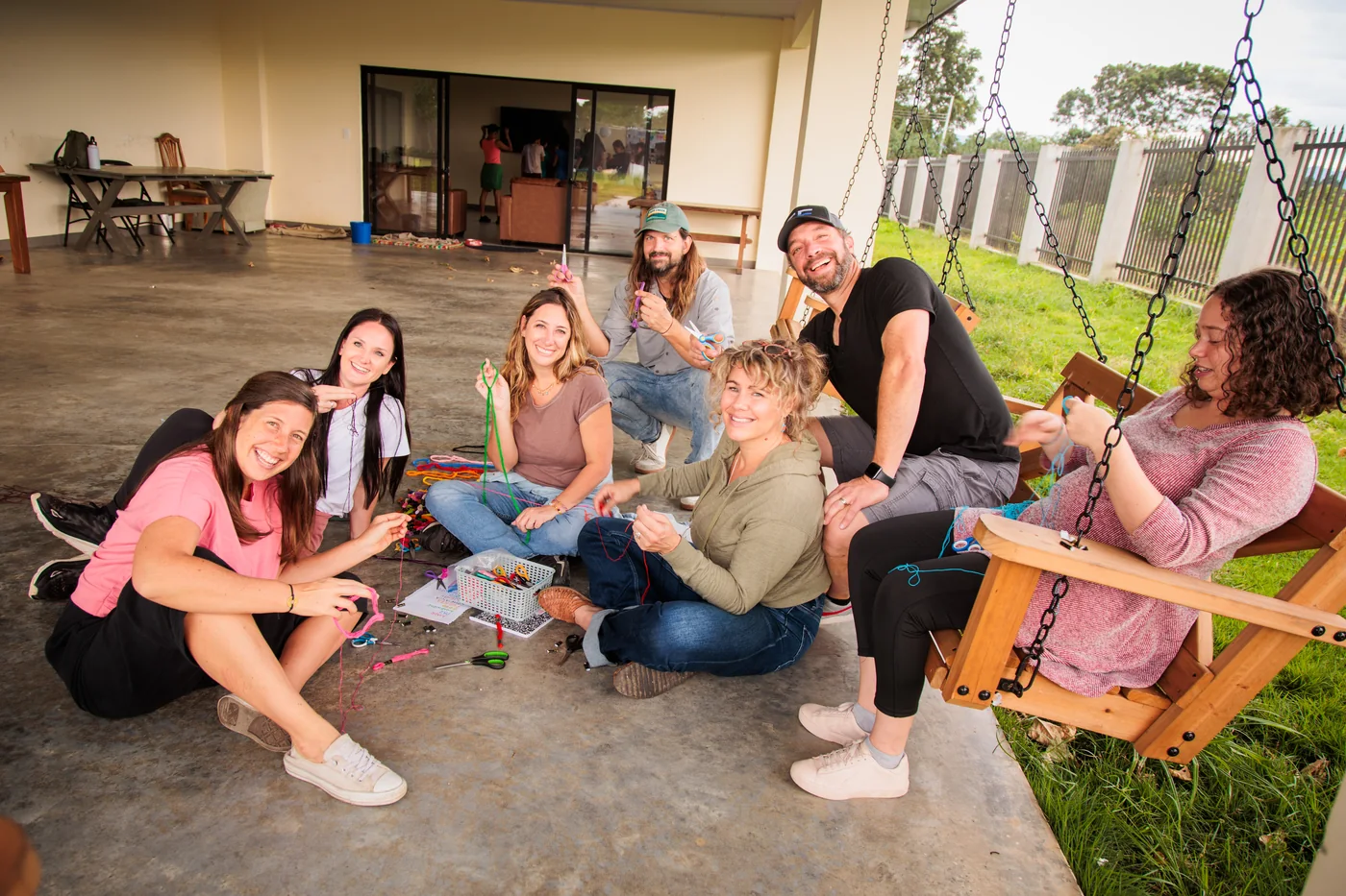 A group sitting together on the floor of an open-sided room in Costa Rica, threading bracelets from a basket of coloured cord, two men among them, everyone looking up and smiling.