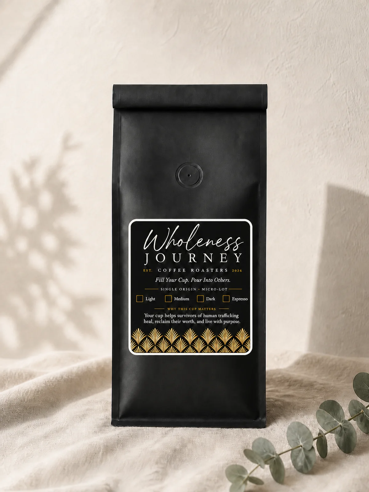 A matte black Wholeness Journey Coffee Roasters bag standing on linen against a plaster wall, its label black with gold fan detail, a eucalyptus sprig beside it.